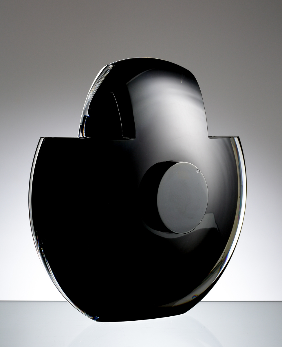 Design: Helen Krantz. Rund och flat skulptur i svart underfång med en större "knapp" på var sida. Vasens övre del har två 90-gradiga utsågningar.