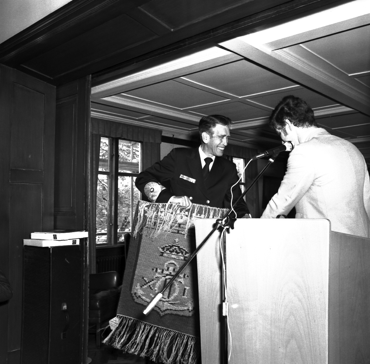 Bilder tagna vid sjösättningsmiddagen i samband med sjösättningen av minfartyget Carlskrona. Viceamiral Rudberg, klädd i uniform, ler och håller en väv i handen. Bredvid honom står en man vid ett podium med en mikrofon framför.