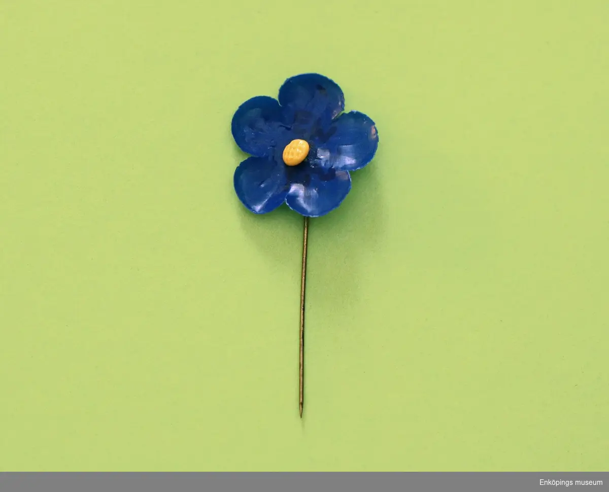 Majblomma från 1923.
Blomman är gjord av marinblå celluloid och har fem blad i ett lager och en gul mittknapp, även denna av celluloid. 
Det som håller blomman samman är en nål av mässing.