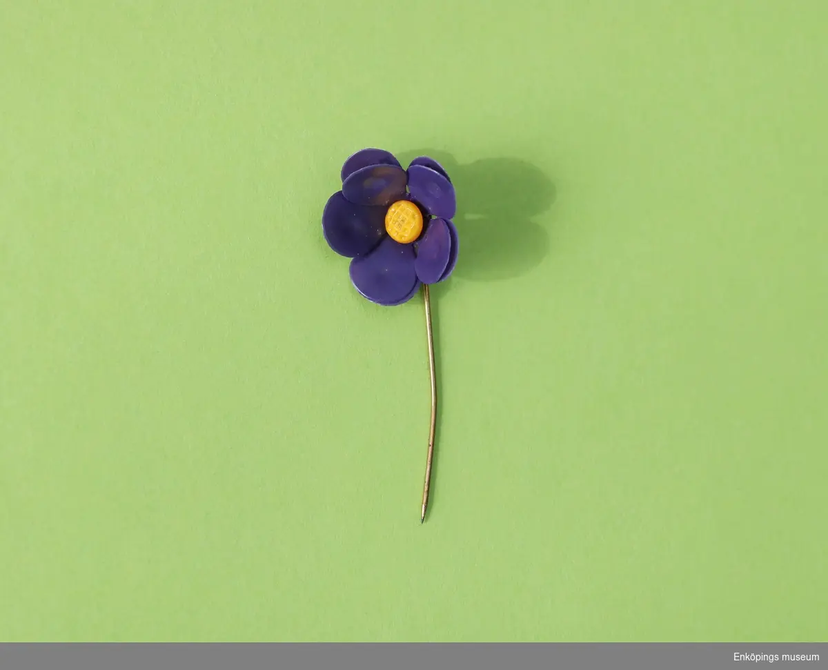 Majblomma från år 1912.
Blomman är gjord av mörklila celluloid och har fem blad i dubbla lager och en gul mittknapp, även denna gjord av celluloid. 
Det som håller blomman samman är en nål av mässing.