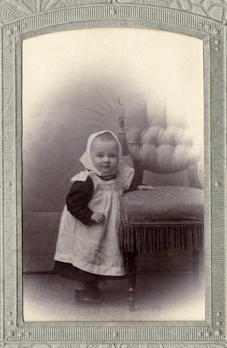 En liten flicka i kolt, förkläde och sjalett som håller sig i en stoppad stol med frans. 
Helfigur. Ateljéfoto.

Fotografens dotter.