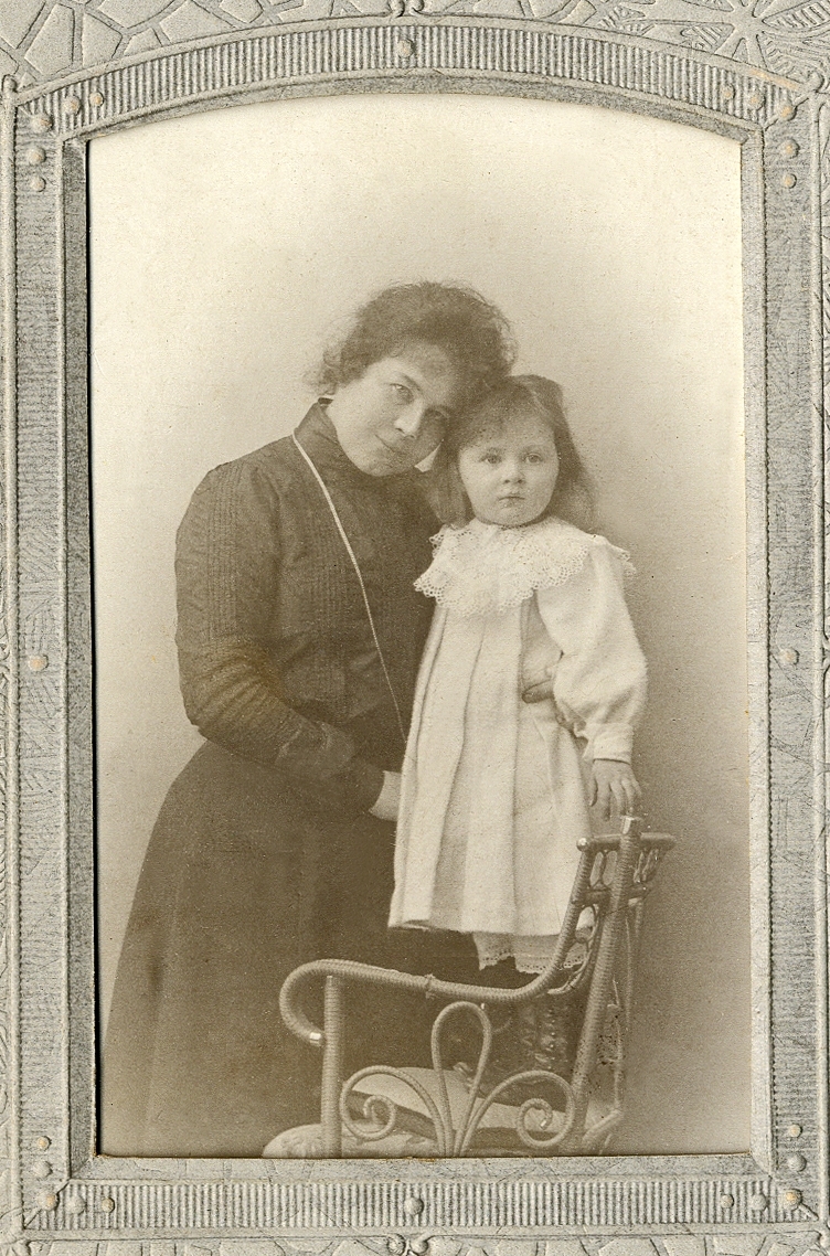En kvinna i mörk klänning med hög krage och en liten flicka i ljus klänning, som står på en rottingstol. 
Knäbild. Ateljéfoto.

Fotografen med dotter.