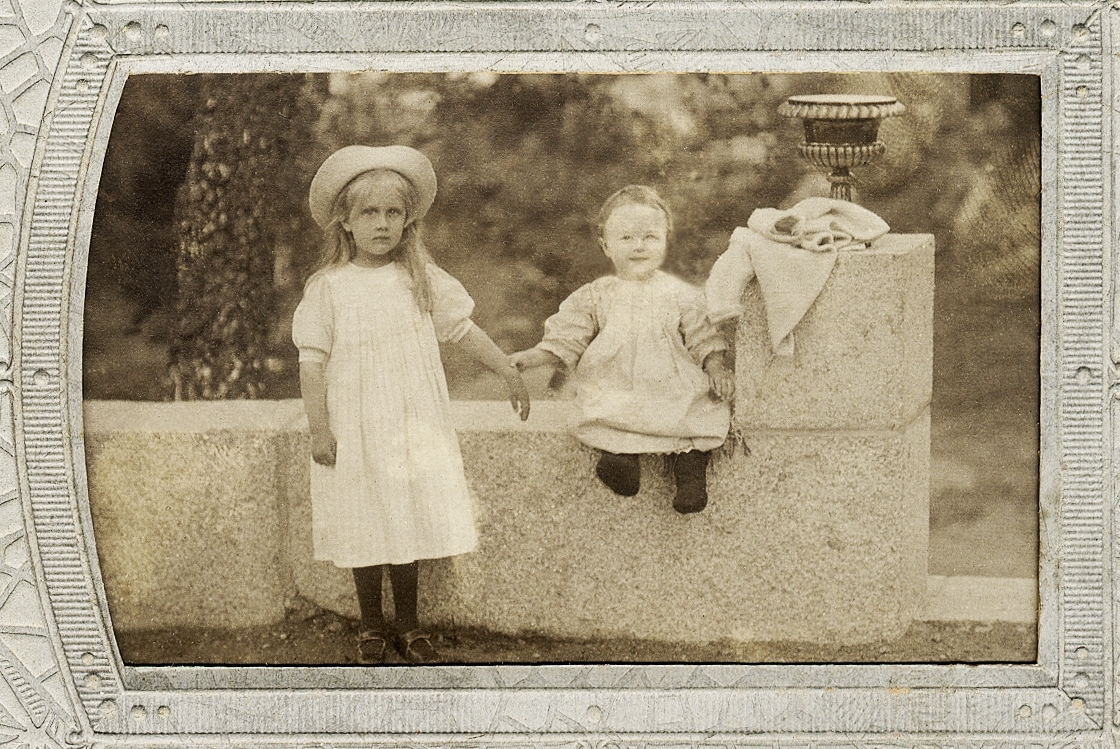 En flicka i klänning, förkläde och stråhatt tillsammans med en liten flicka i kolt, som sitter på en stenmur.
