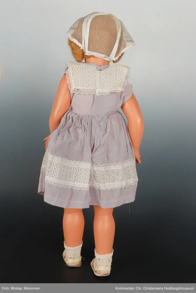 Dukke med blondt hår, kysehatt, syrinfarget kjole, hvite halvsokker og plastsandaler. 