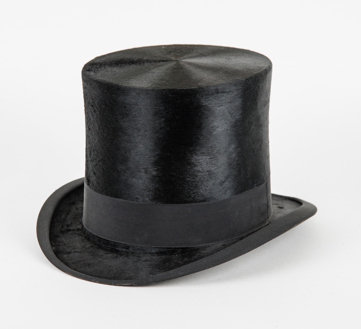 Hatt, høg svart flosshatt i silkefloss. Ripsband rundt pullen og bremen. Skinnband på innsida.
