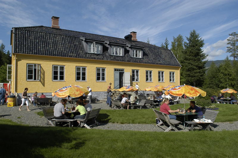 Bildet viser uteområdet bak Lysaker gjestgiveri. Folk sitter på benker under parasoller og spiser mat.