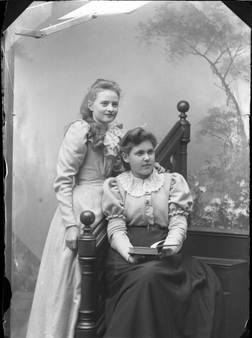 Två unga kvinnor intill varandra. Ester Grönberg står till vänster och har sin hand på axel på en okänd kvinna som sitter i trappa med en bok i knät.