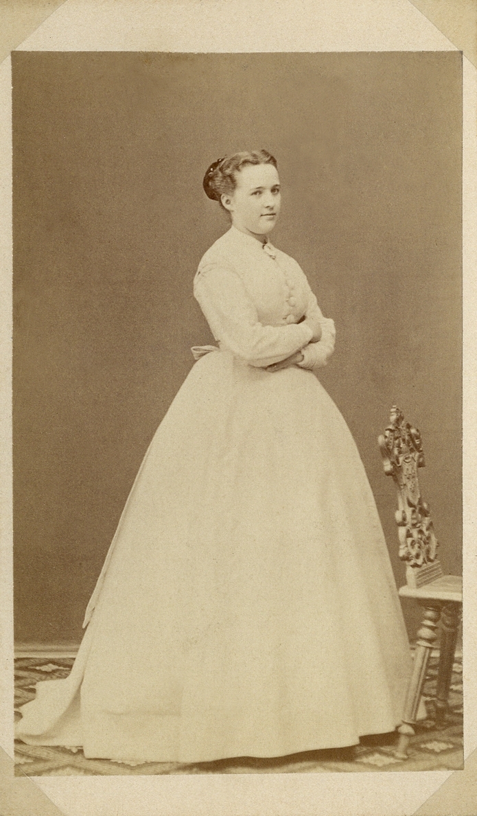 Porträttfoto av en ung kvinna i ljus klänning med liten vit krage. Framför henne syns en snidad stol. 
Helfigur, halvprofil. Ateljéfoto.