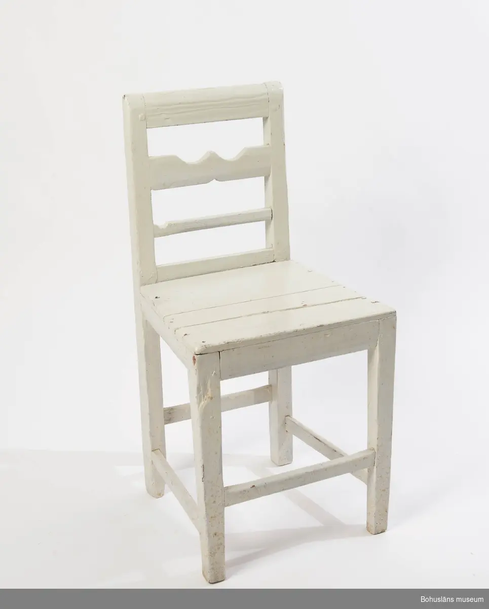 Vitmålad enkel stol med slät sits och nertill fyra tvärslåar på olika höjd.
Ryggstöd med två  tvärslåar, den översta en sk. "krusabete", en dekorativ tilltäljd utsirning på mittslån.
