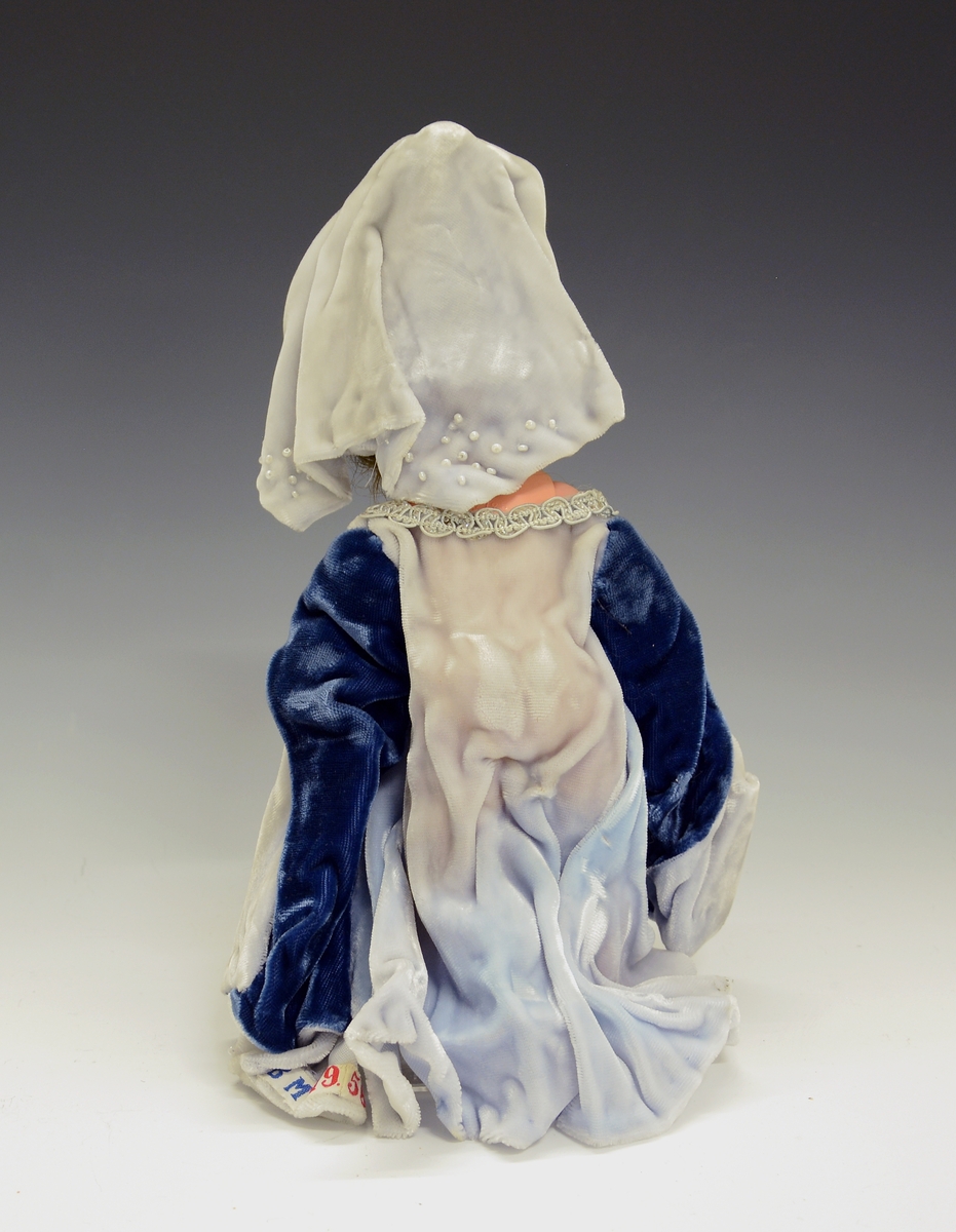 Dukke av plast i blå kjole. Dokka skal forestille gotisk middelalder ca. 1200-1480 og den sitter på en hvit hest med rødt seletøy.