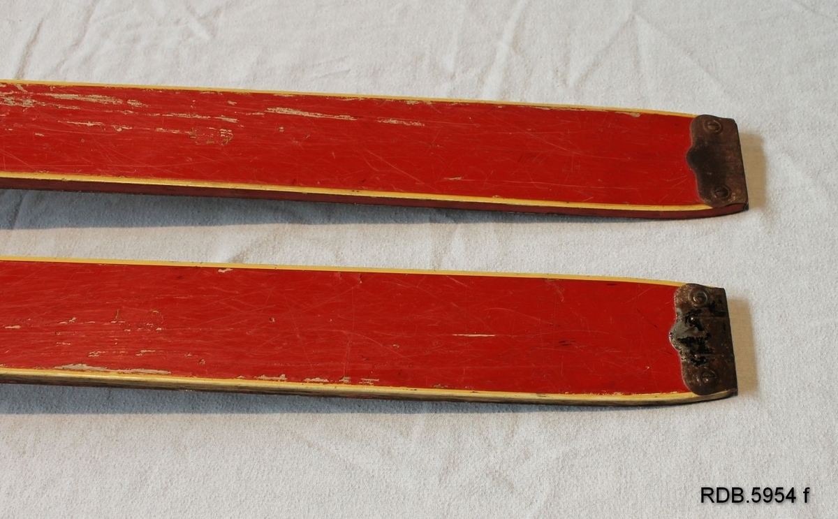 Et par røde, laminerte barne-treski med stålkanter og stålbeslag bak. På venstre ski er det noe malingssøl. Kandaharbinding og blå fotplate.