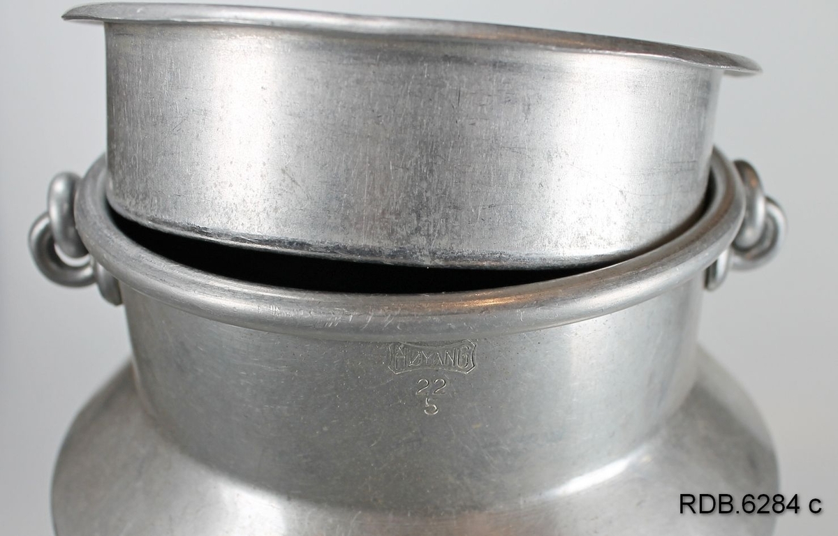 Sølvfarget aluminiumsspann med hals, hengslet hank og løst lokk med håndtak.