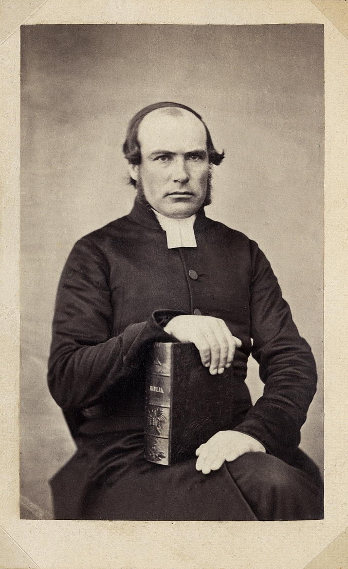Porträttfoto av en man i prästdräkt m.m. Han håller en Bibel mot ena knäet. 
Knäbild, halvprofil. Ateljéfoto.
