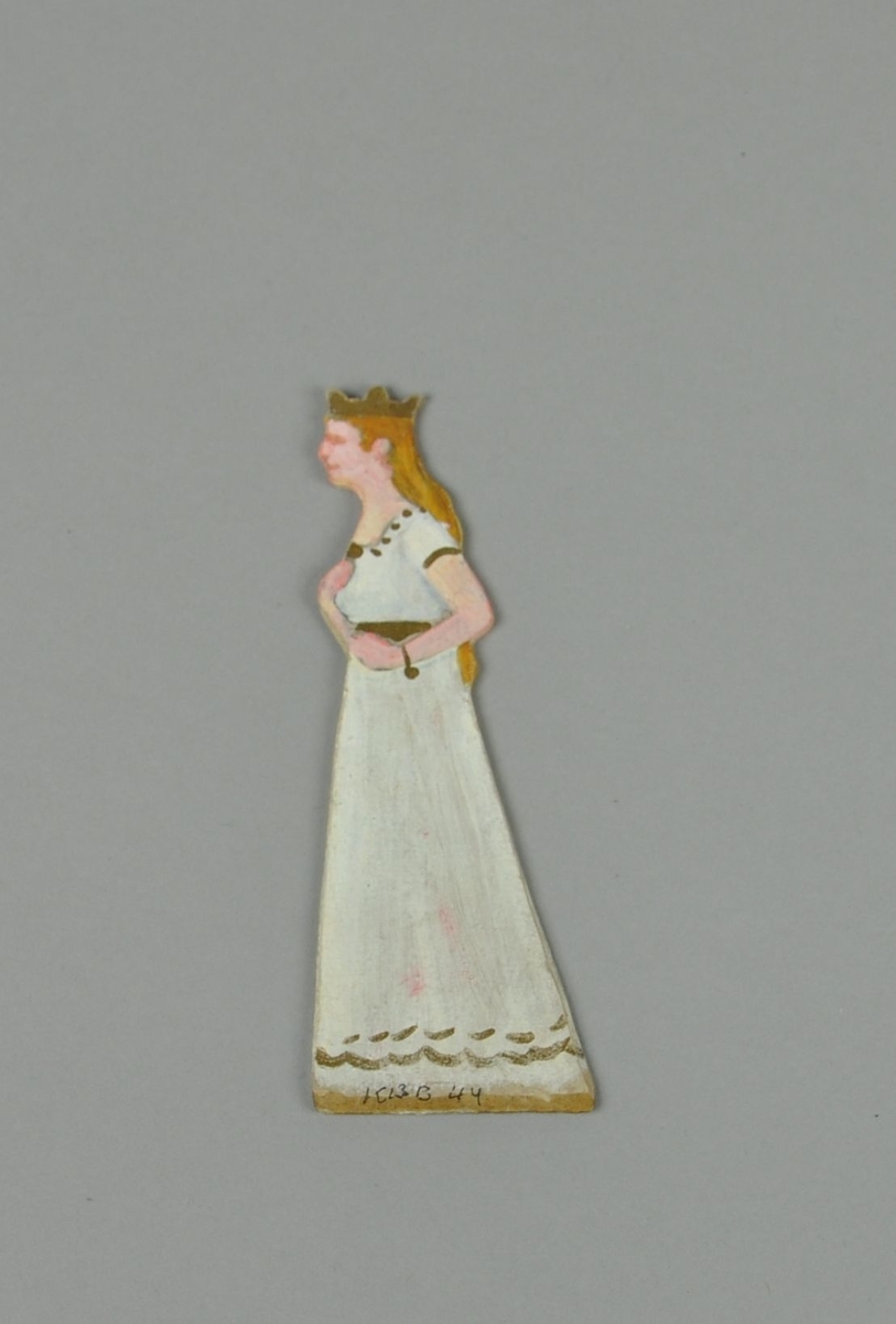 Figur av papp. Figuren forestiller stående dame i sideprofil, med krone og hvit kjole. Figuren er malt på begge sider.
