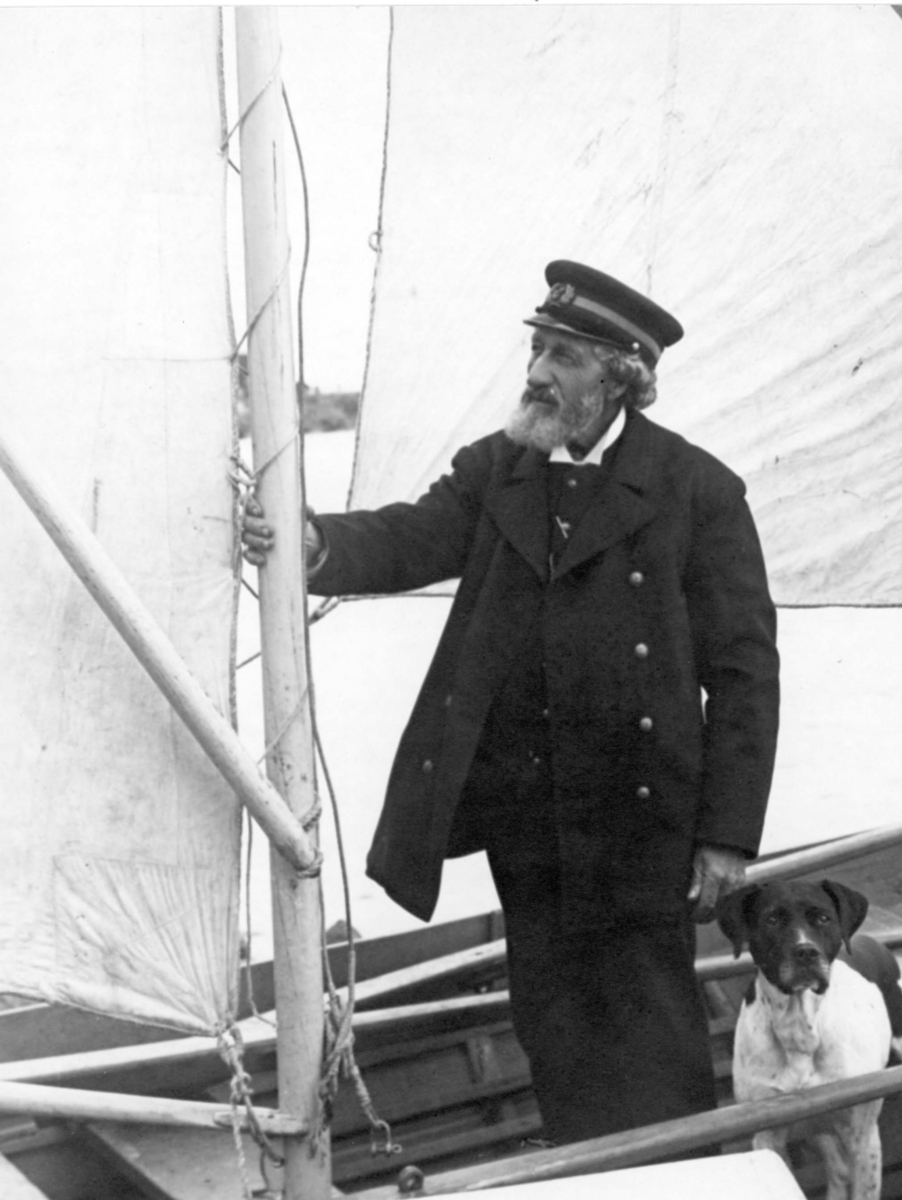 Kapten Zetterberg, i uniform, står i en liten båt med hissade segel. Han håller sig i masten och har en hund vid sin sida.