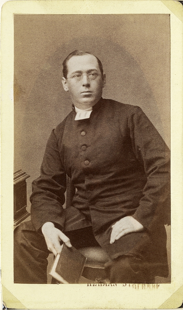 Foto av en man i glasögon, klädd i prästrock och prästkrage m.m. Han håller en bok i ena handen. 
Knäbild, halvprofil. Ateljéfoto.