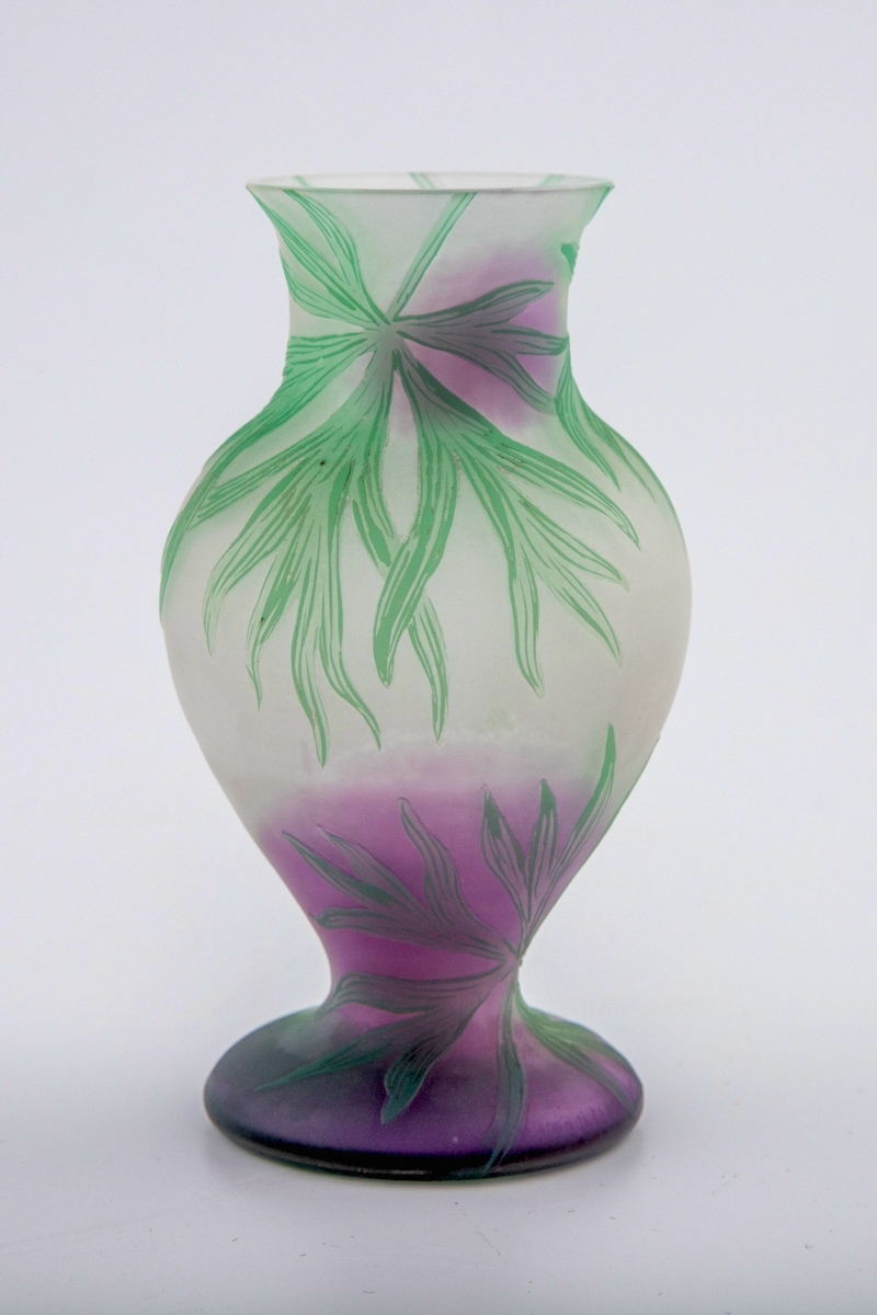 Balusterformet vase i farget glass. Øvre del av vasen er utført i halvgjennomskinnelig melkehvitt glass, mens nedre del er lillafarget. Korpus er dekorert med grønnfarget bladverk i overfangsteknikk.