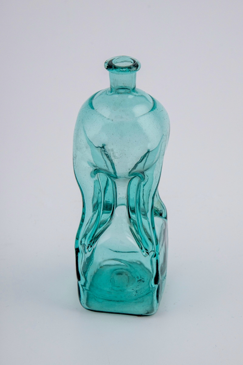 Lav klukkflaske i gjennomskinnelig turkisfarget glass. Timeglass-lignende utforming, hvor den kvadratiske over- og underdelen er forbundet med et tynt rør. Lav hals med glatt kantring, samt puntemerke på undersiden.