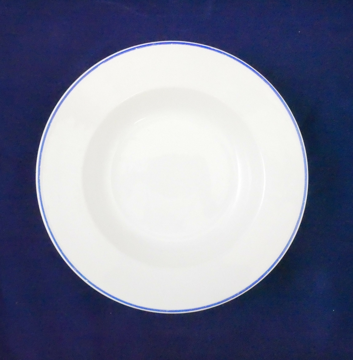 Hvit dyp tallerken i porselen. Rundt kanten er det en blå linje. 