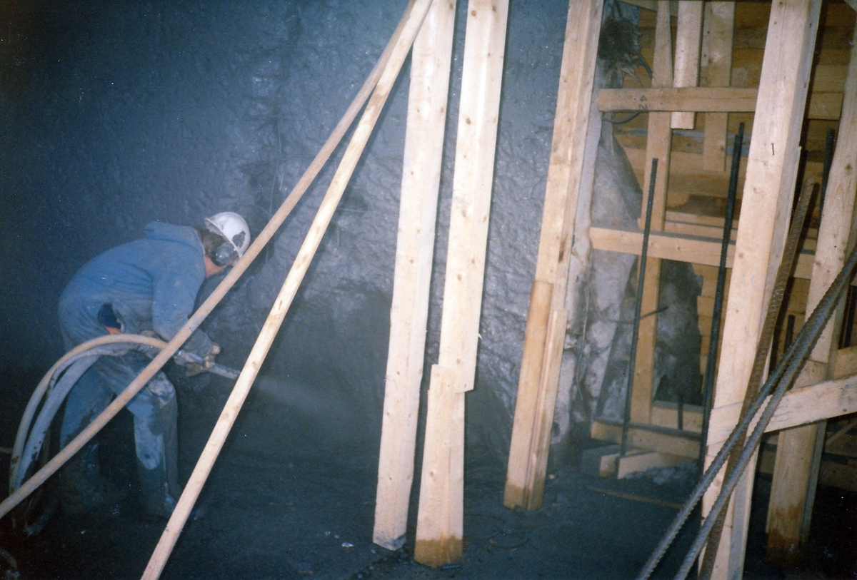 Anleggsarbeid for kraftverket på Kistefoss. Mann med kjeledress og hjelm spyler betongvegg. Forskallingskonstruksjon i treverk til høyre i fotografi.