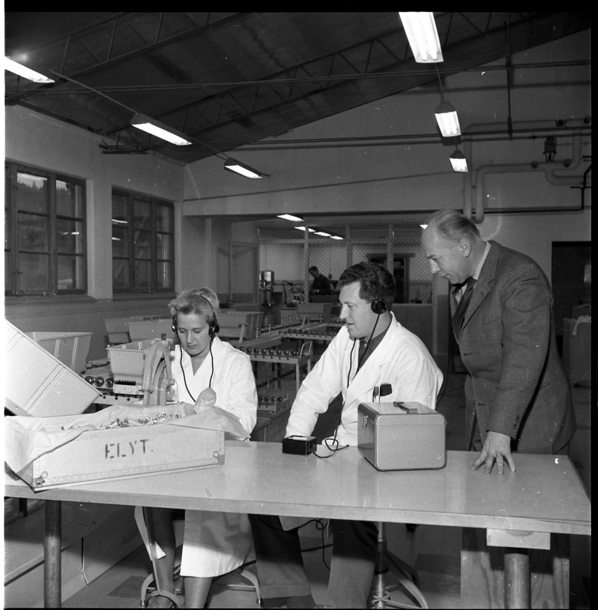 En kvinna och en man i vit arbetsrock sitter vid en arbetsstation på Rifa-fabriken i Gränna. De har öronskydd/hörlurar. Till höger står en man något lutad över dem. De skrattar.
De är från vänster Inga Johansson, Göran Andersson och G Eklund.