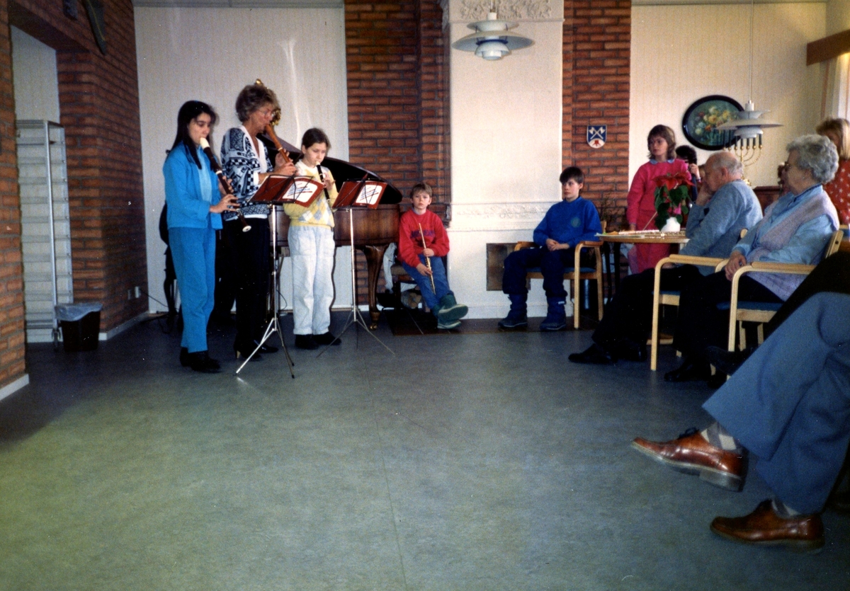 Musikskolan är på besök i Brattåsgårdens matsal på Streteredsvägen 5, cirka 1986. Gruppen består av lärare och barn som spelar träblåsinstrument (blockflöjt).