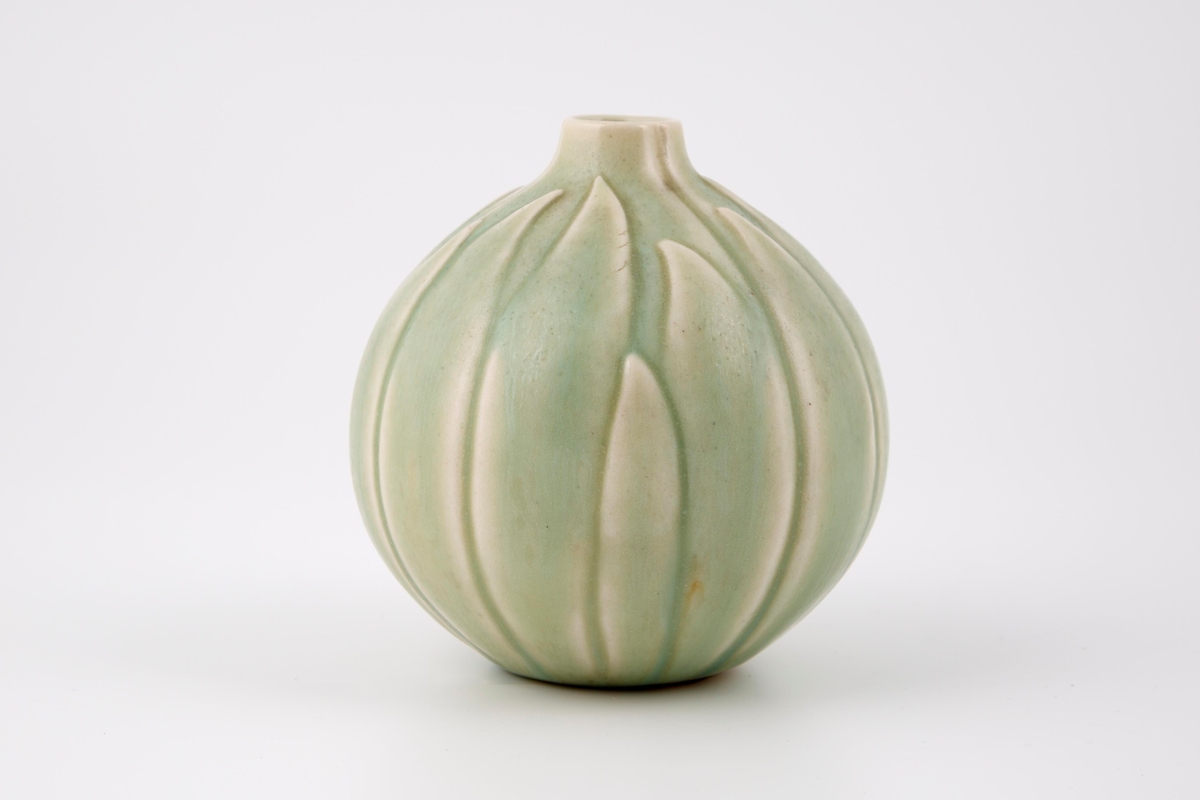 Liten, kuleformet vase med forhøyet, trang munning. Vasen er utformet som en knopp med blader i relieff. Retningen på bladene er fra bunnen og opp.  Den har en blek grønn glasur som kun dekker deler av vasen og dermed eksponerer godset og gir relieffet konturer og dybdeeffekt.