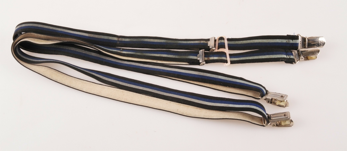 Selve selen er delt i to like deler, som består av et vevd elastisk "bånd", der gummistrikken er skjult i ripsveving med striper i svart, lys blå, blå og svart på retta, gråhvit på vranga. Hele lengden av "båndet" er ca 125 cm. I ene enden er det sydd fast en metallklype. Tilsvarende klype ligger i bukten av "båndet". Andre enden av "båndet" har en spenne festet fast og med spalte der "båndet" på "tilbakevei" er tredd gjennom. Her er det mulig å åpne spenna for regulering. De to seledelene trees i hver sin spalte i en rosa plast "spenne". Den er defekt.