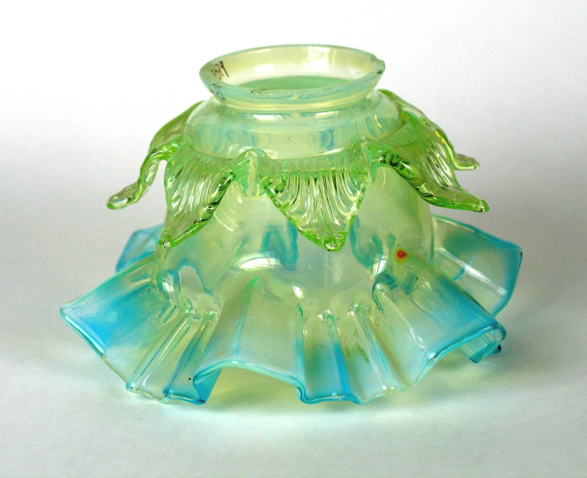 Lampeskjerm i glass med turkis bølget kant nederst og en lys grønn bladformet kant øverst. Trearmet metallfeste med skrufeste. Løst.