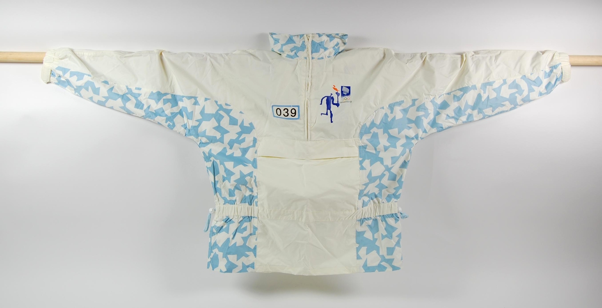 Overtrekksdress med hovedfargene hvit og blå. Logo for de olympiske vinterleker på Lillehammer i 1994 og fakkelstafetten på brystet og ryggen på jakken. Jakka har hette som kan foldes inn i kragen, glidelås på brystet og en stor lomme på magen.