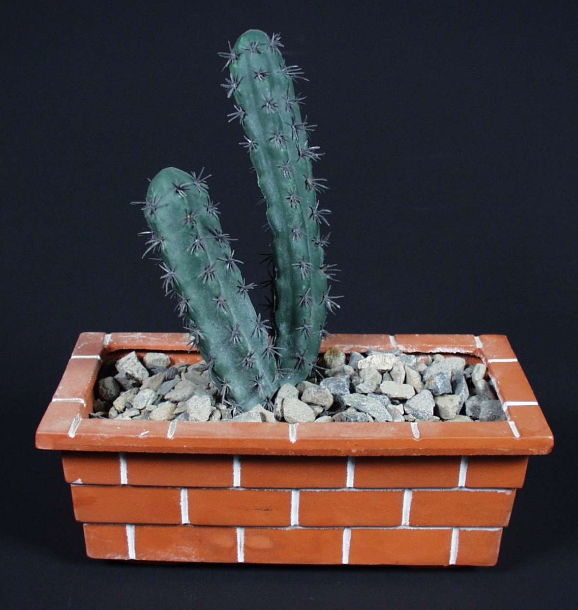 Firkantet blomsterpotte i keramikk med mønster som ligner teglsteinsmur. Kunstig kaktus med stein rundt.