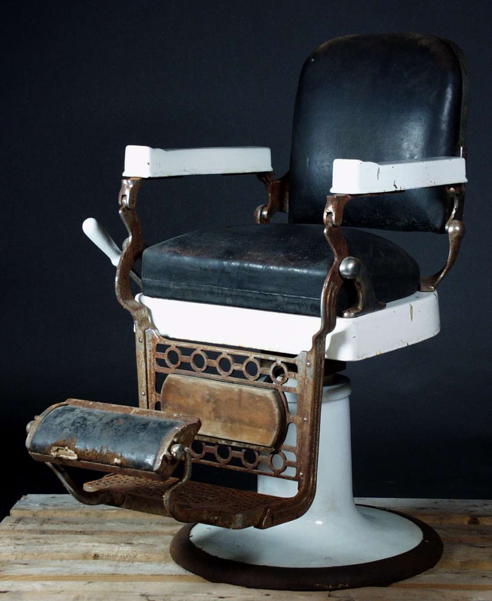 En barberstol i metall og støpejern. Rygg og sete er lærtrukket. Den har hvite armlenere og understell.
På understellet er det en sveiv hvor sete kan heises opp eller ned. Fotsøylen er i støpejern og lær. Den har to trinn. 
