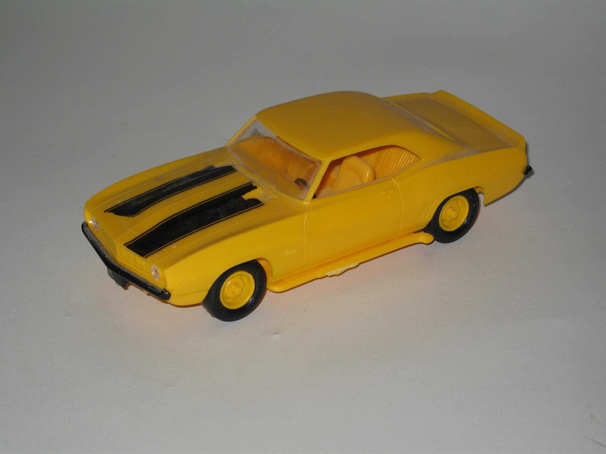 Leketøysbil bestående av karosseri og understell av gul plast påsatt klarplast vinduer foran og bak, svarte støtfangere foran og bak, fire svarte dekk med gule felger og to klarplast frontlykter. Svarte striper limt på panseret og blå bilskilt med gul tekst.
Bilen er en modell av Chevrolet Camaro.