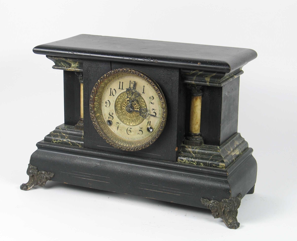 Klokkekasse med kvadratisk midtfelt med rund tallskive, inntrukte sidefelter med opprinnelig to halvsøyler på hver side, de ytterste senere fjernet. Topplate med avrundet kant, profilert sokkel og på hørnene påsatt mønstrede messingbein. Svartmalt og litt bruk av grønn marmorering. Topplaten er flat, og har gitt plass til eventuelle løse figurer på toppen. På sidene av klokkekassen har det også vært dekorative messingornamenter.