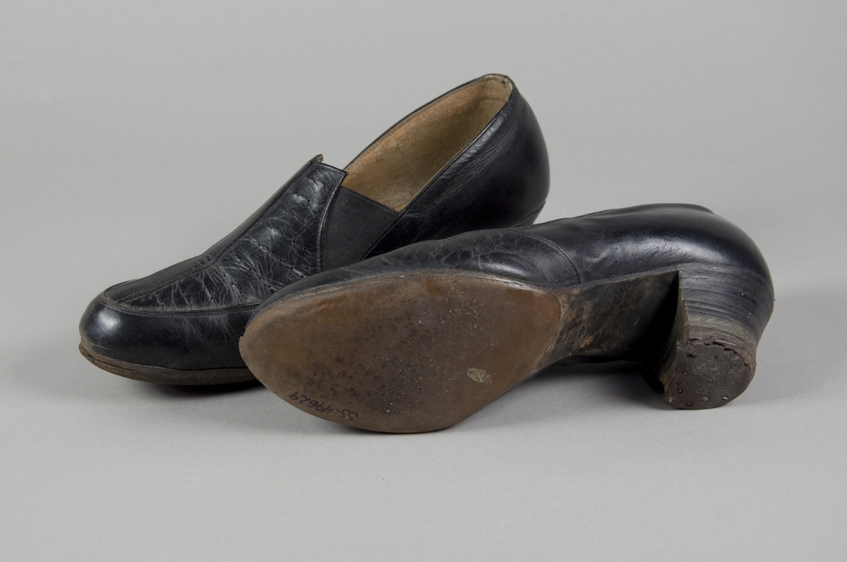 Sorte skinnsko med relativ høy hæl. Vriststykket har hulldekor og strikk på hver side.  Skoene har helfôr og er halvsålet.