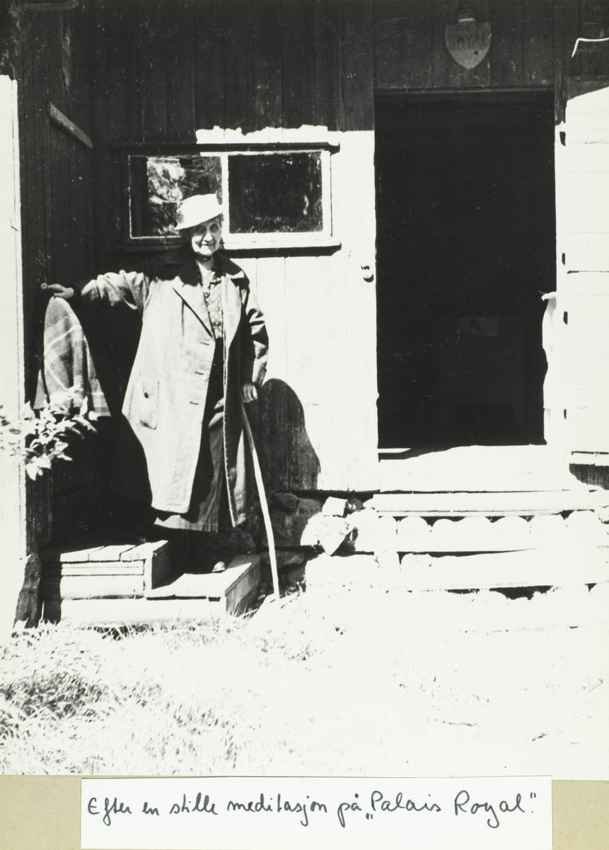En eldre dame med stokk, hatt og kåpe utenfor hus.