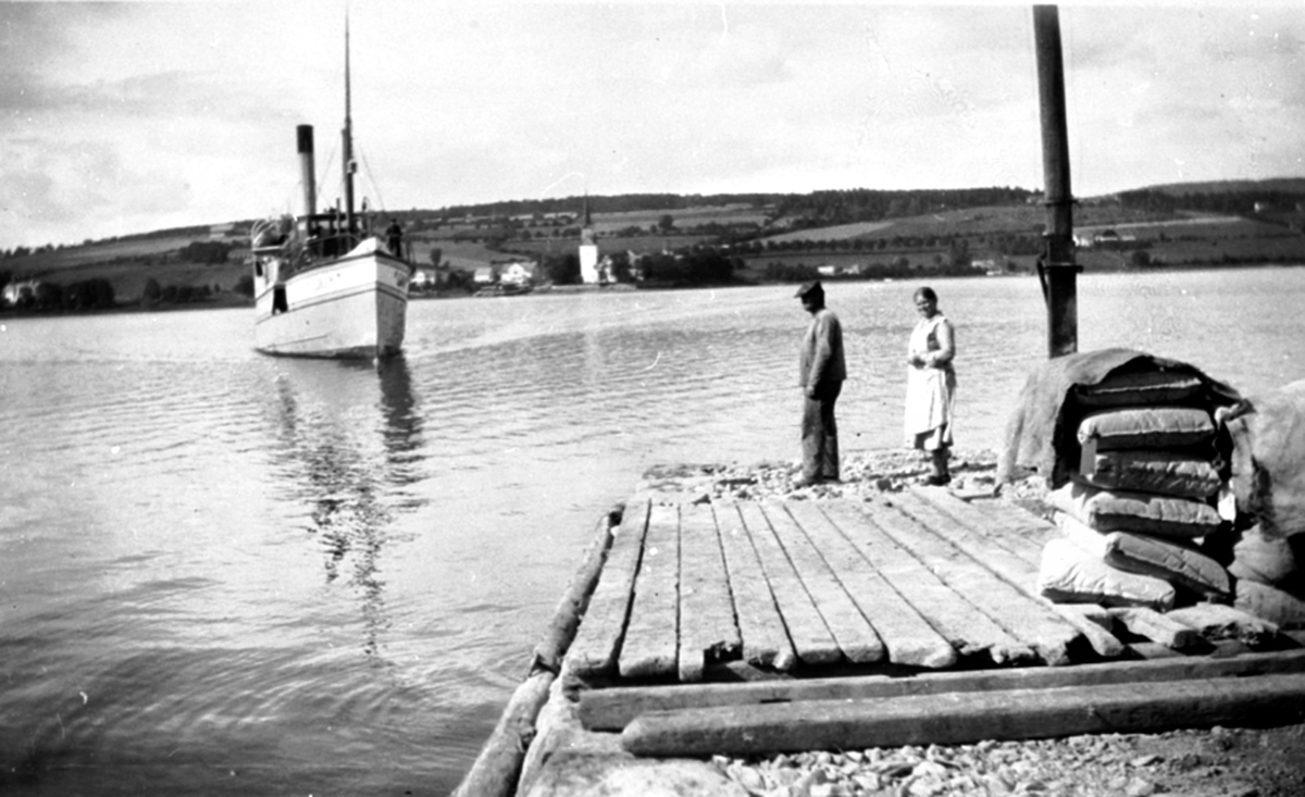 Mjøsbåten D.S. Gjøvik på vei inn til Sundsbrygga. Karl Sund og Helga Kleven tar i mot. Karl og Helga bodde i Sundgård, Helgøya. Hun betjente telefonsentralen.Tingnes og Nes kirke i bakgrunn.