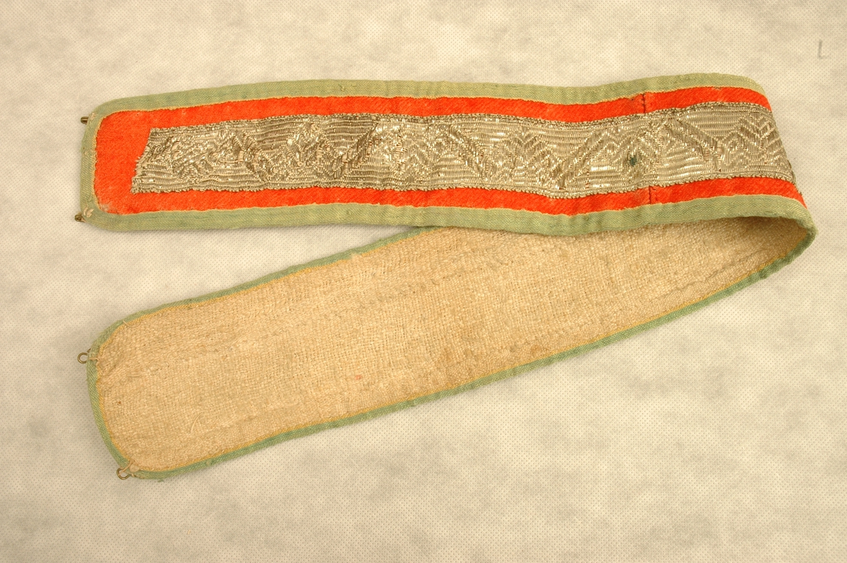 Belte av rødt ullstoff kantet med grønne linband. Midt på beltet er sydd på et 5 cm bredt vevd metallband. Metall hemper og hekter til lukking i hver ende. Beltet er fóret med grovt linstoff.