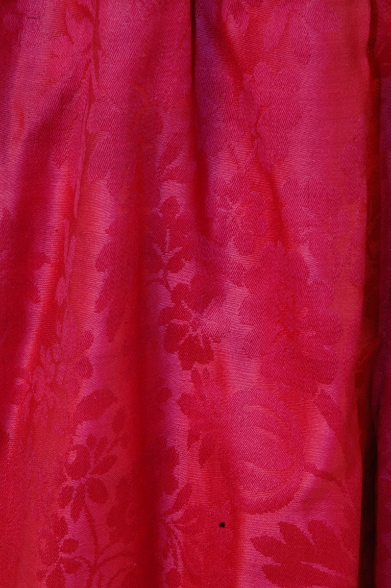 Rød stakk av glanset ulldamask. Foldet med motfold midt bak, lukking med hekte i venstre side. Linning av rød bomullssateng.