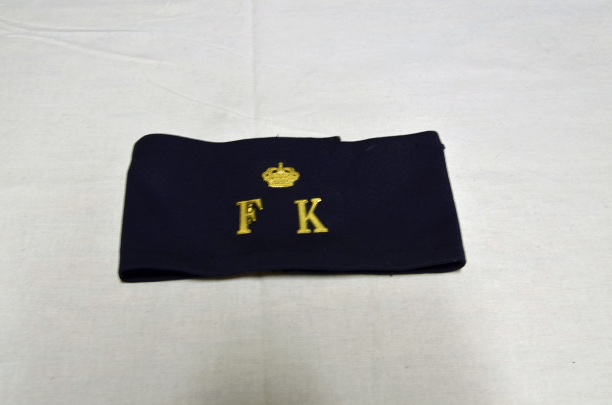 Armbindel i tyg med initialerna F, K samt en krona i guldfärgad metall.