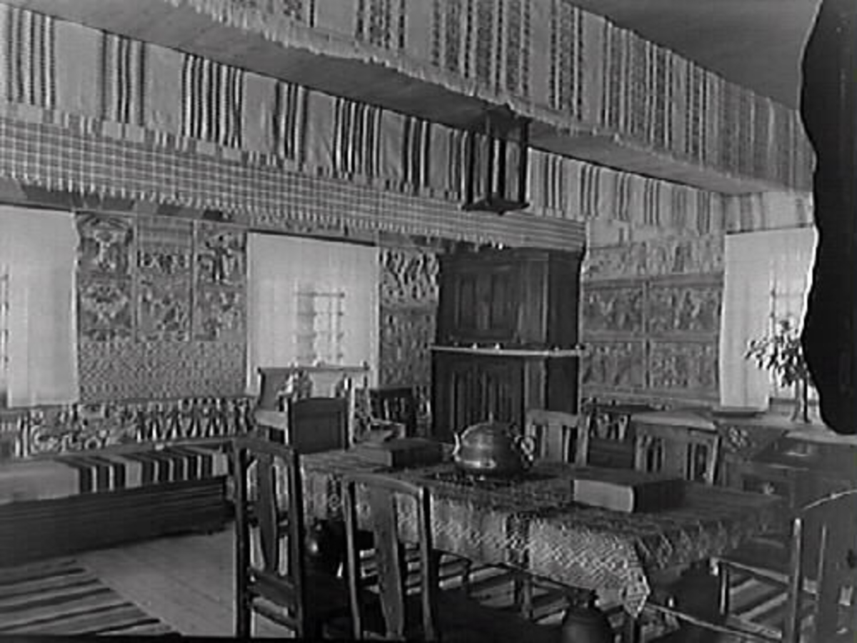 Interiör av bålastugan, Bexellska ryggåsstugan, som är dragen med halländska bonadsmålningar och hängkläden. Matbord i mitten och ett hörnskåp.