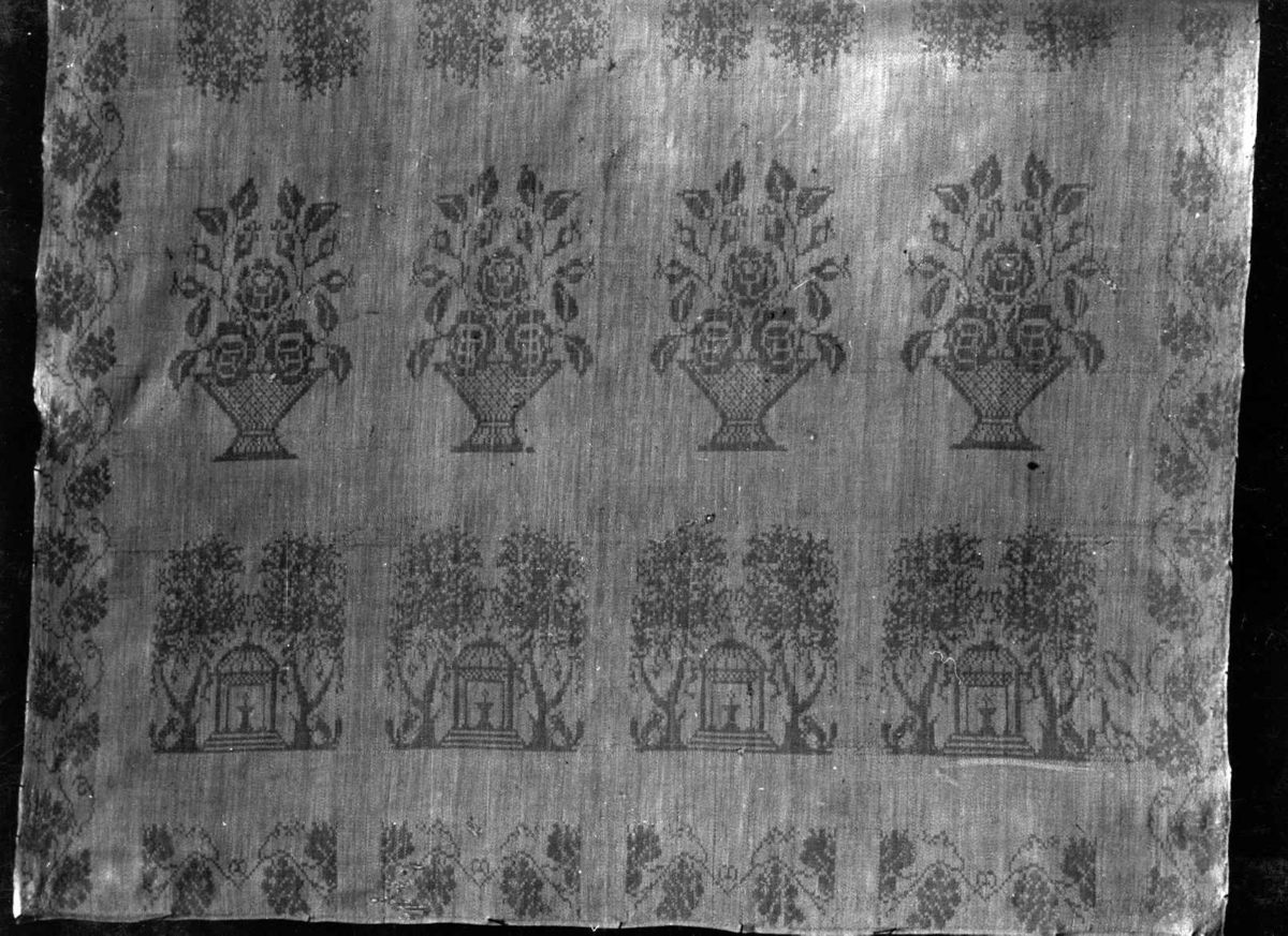Hvit serviett med avvekslende rekker av lysthus, trær og blomsterkurver, rankebord. Fra 1835, muligens Røyken, Buskerud. Norsk hjemmevevet damask.