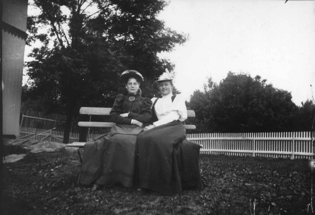 Drakt 1908-1910. To kvinner sittende på en benk i en hage.  Bærer hatter.