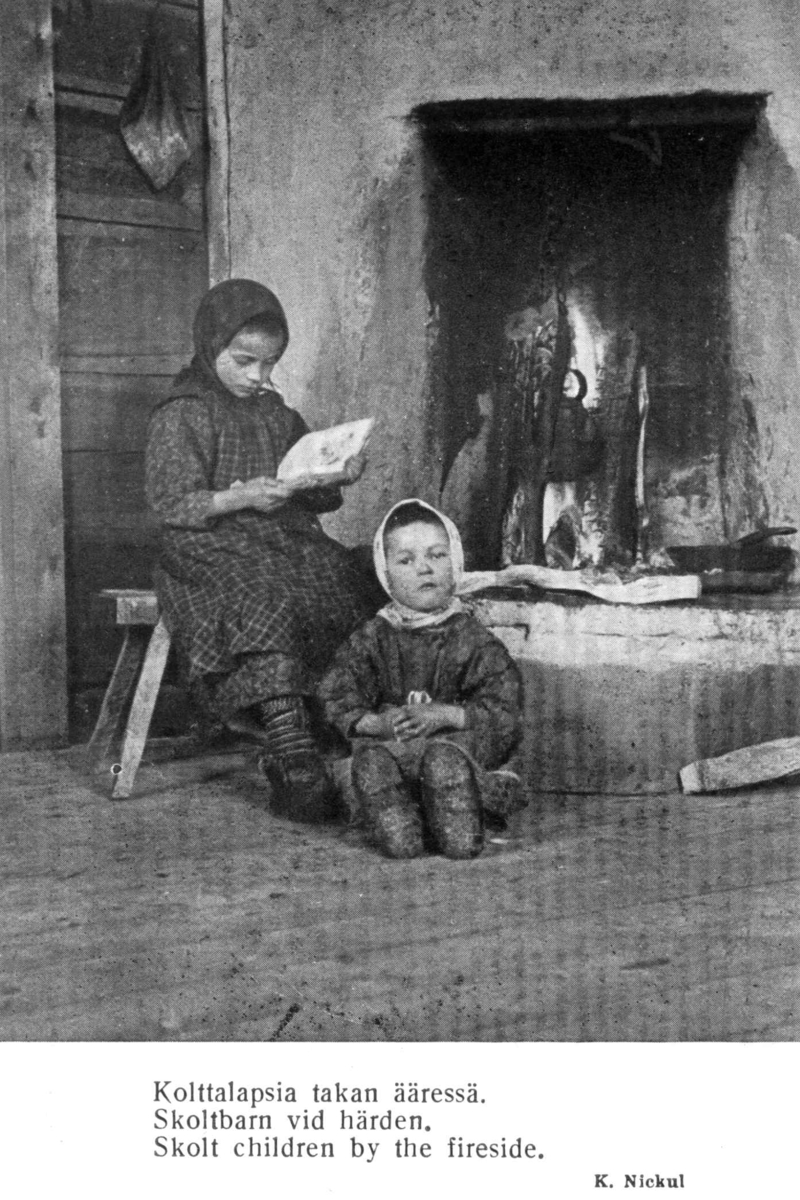 To skoltebarn sitter ved peisen, jenta leser en bok.