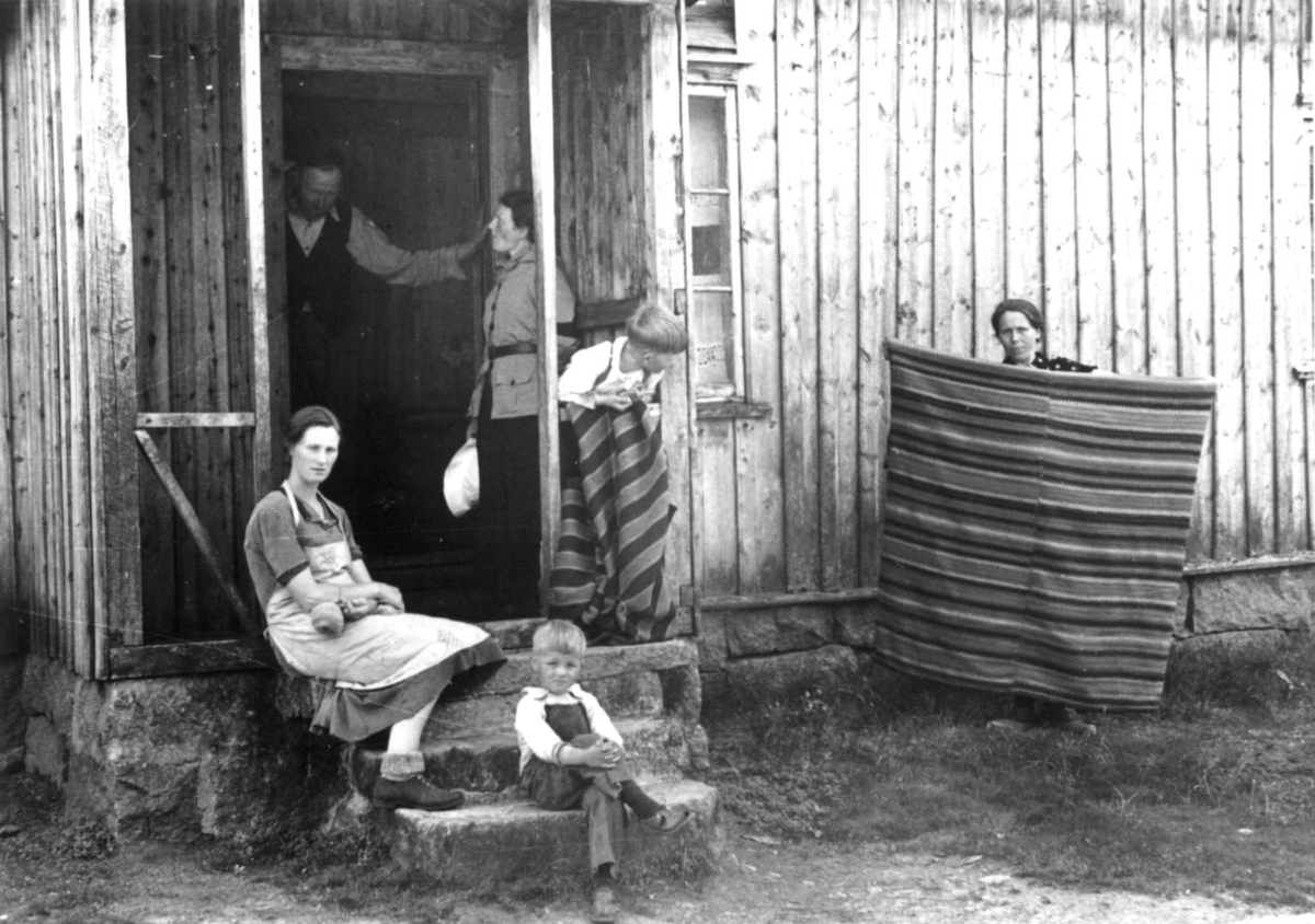 Anstein Åvitsland med flere samlet utenfor inngangspartiet til huset hans. Datteren viser frem et gammelt sengeteppe. Fjotland, Kvinesdal, 1941.