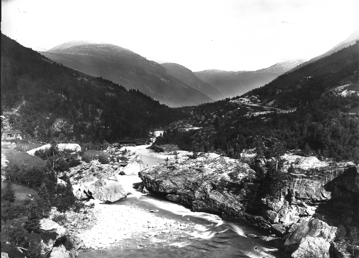 Landskap, ukjent sted. Skogkledd dalområde med elveparti, veiparti i bakgrunnen t.h. Del av Den Norske Turistforenings samling landskapsbilder, de fleste tatt av fotografene Brødrene Brunskow.