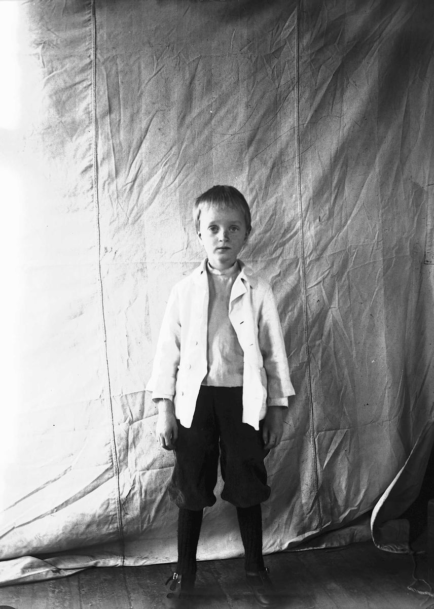 Portrett av gutt, fotografert mot opphengt bakteppe, ukjent sted. Antatt en av barna i familen Axel Q. Wiborg.