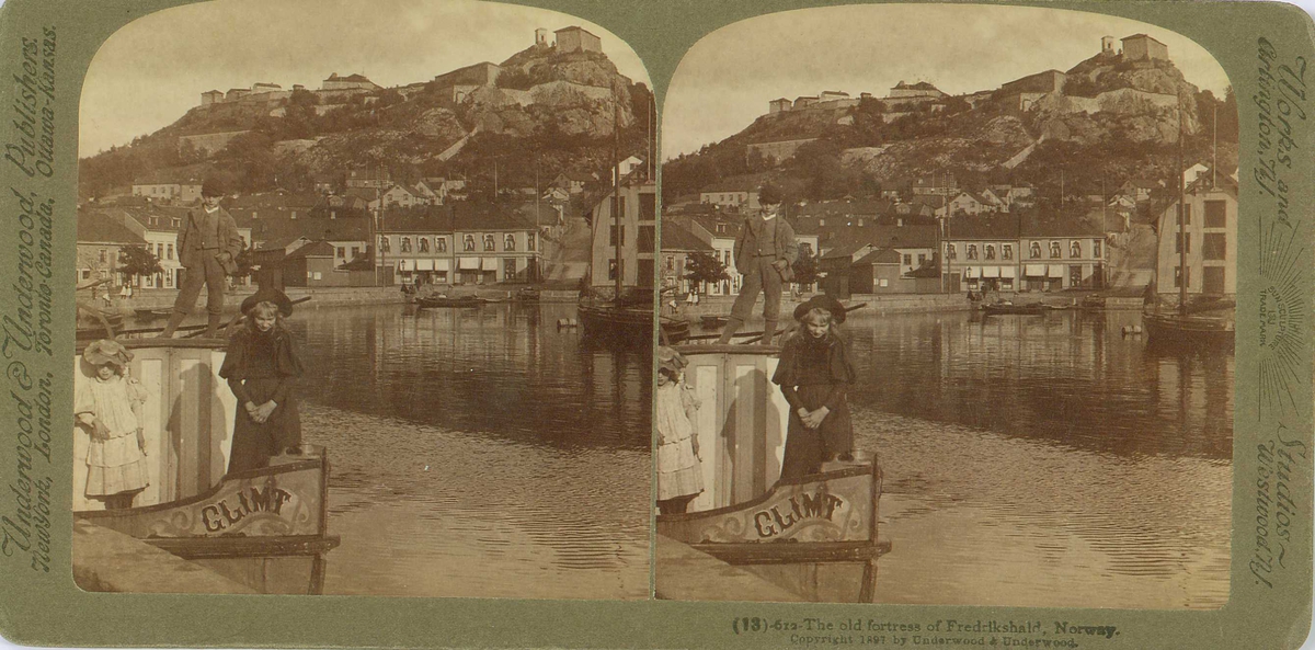Halden med Fredriksten festning. I forgrunnen barn i båten "Glimt" ved kai. Stereofotografi utgitt av firmaet Underwood&Underwood.