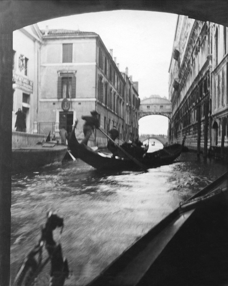 To gondolister manøvrerer gondolene igjennom kanalene i Venezia. Sukkenes bru i bakgrunnen. Robsahm og Lund.