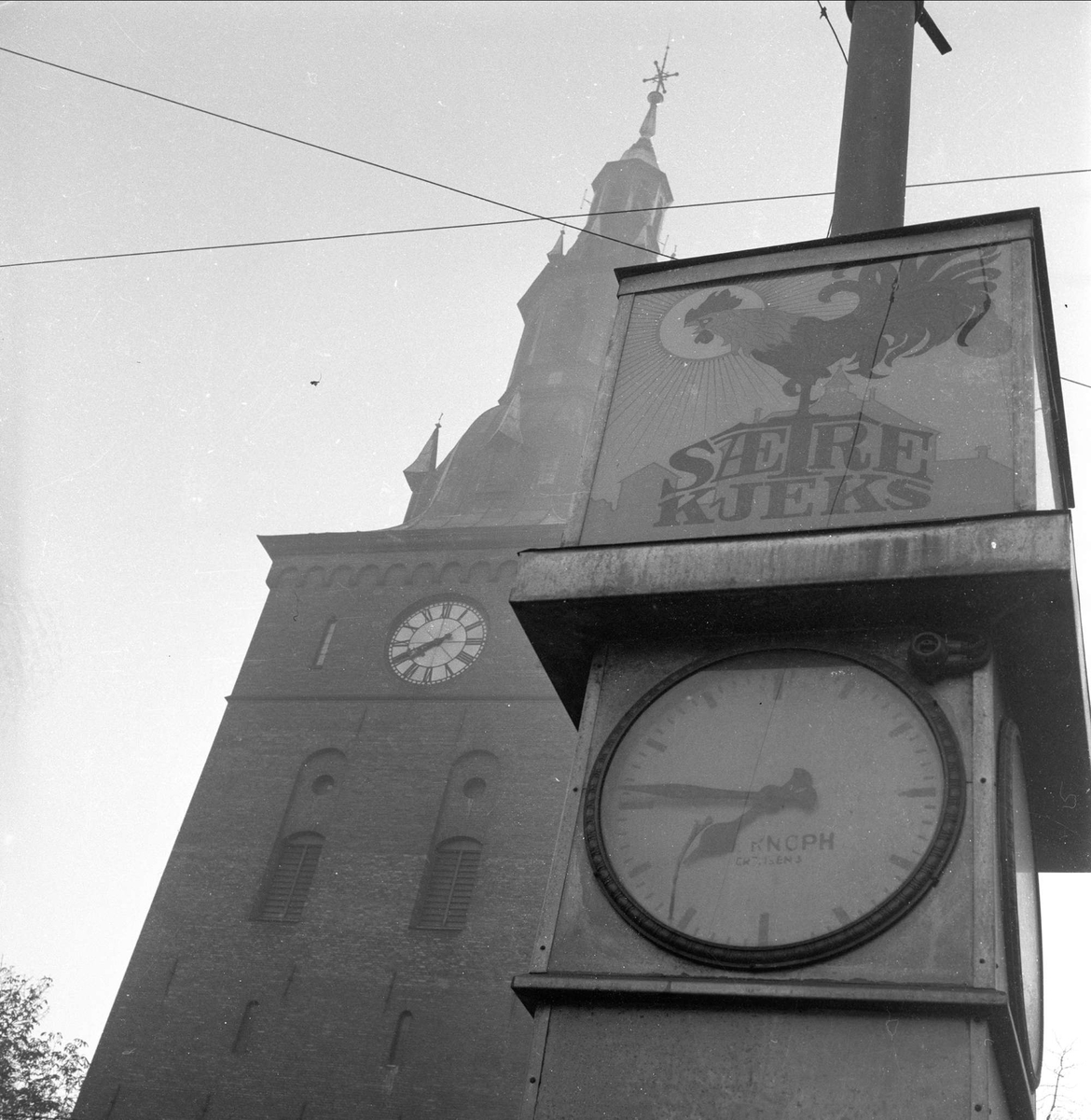 Oslo, 15.10.1957. Domkirken bak stolpe med klokke og reklame.